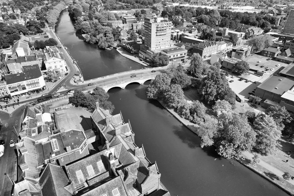 An aerial black-and-white photograph of Upminster Bridge showing the River Ingrebourne flowing beneath a white stone arch bridge. The river is bordered by lush, leafy trees on both sides, with some tr