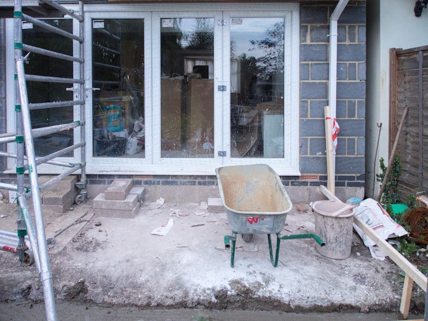 A residential garden undergoing landscaping work with a partially paved patio area in front of a large glass sliding door. The patio surface appears to be in the process of being laid, with exposed so